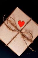 handgemaakte geschenkdoos verpakt in ambachtelijk papier met rood houten hart, touw en strik op zwarte achtergrond. bovenaanzicht, plat gelegd foto
