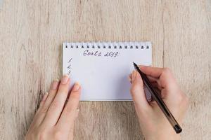 jonge vrouw handen houden geopende notebook pagina's met zwarte pen op houten tafel. doelen 2019 lijst. foto