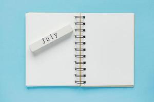 schoon spiraalvormig notitieboek voor notities en berichten en juli houten kalenderbalk op blauwe achtergrond. minimaal zakelijk plat leggen foto
