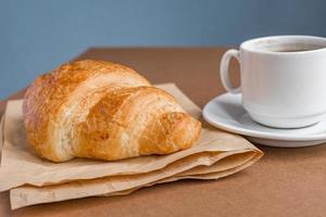 smakelijk ontbijt. Franse croissant geserveerd op ambachtelijk papier en kopje zwarte koffie of espresso op bruine achtergrond. foto
