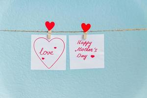 rode liefde harten pin opknoping op natuurlijke koord tegen blauwe achtergrond. gelukkige moederdag inscriptie op stuk papier. foto