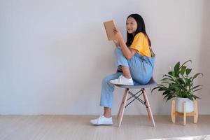 de meisje zittend Aan een stoel Holding een boek in een minimaal kamer. foto