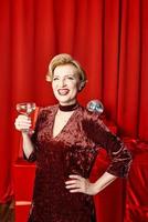 volwassen elegant elegant vrouw met glas van sprankelend wijn met presenteert Aan rood achtergrond. partij, mode, viering, anti leeftijd concept foto