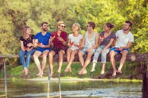 vrienden genieten van watermeloen terwijl zittend Aan de houten brug foto