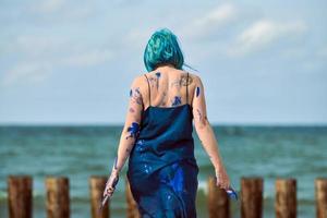 artistieke blauwharige vrouw performancekunstenaar besmeurd met blauwe gouacheverf dansend op het strand foto