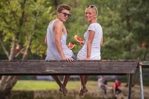 paar genieten van watermeloen terwijl zittend Aan de houten brug foto