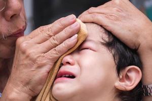dichtbij omhoog van een 1-2 jaar oud Aziatisch gezicht jongen is huilen van pijn in zijn voorhoofd. opa toepassingen ijs verpakt in een kleding naar koel de gewond Oppervlakte. elementair verpleegster beoefenaar. foto