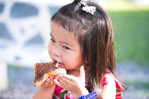 portret beeld kind 3-4 jaren oud. aanbiddelijk kind genieten van heerlijk met chocolade bedekt brood. hongerig kinderen aan het eten snacks Bij park. gedurende zomer of voorjaar tijd. Aziatisch meisjes genieten van voedsel. zoet glimlach foto