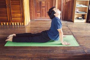 gezond jong Aziatisch Mens aan het doen yoga beoefenen cobra asana in de kamer Bij houten huis. foto