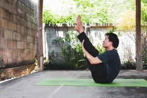 gezond leven concept van jong Aziatisch Mens beoefenen yoga asana paripurna navasana houding of vol boot houding, werk uit en poses Aan een groen yoga mat. buiten oefening in de tuin. gezond levensstijl. foto