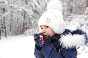 een weinig meisje in de verkoudheid verwarmt haar handen in wanten-ademen warm lucht van haar mond Aan haar handen. winter, wandelen de baby in de Open lucht, de sneeuw. warm kleding, gebreid hoed, vacht POM Pom en kap foto