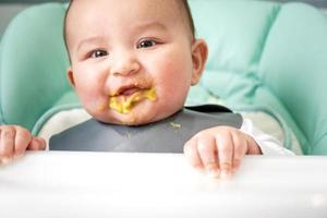 een vies, blij baby Bij de tafel Aan een voeden stoel kreeg zijn mond vuil in groente puree. invoering van complementair voedsel, de kind houdt naar eten. portret detailopname foto