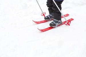 kindervoeten in rode plastic ski's met stokken gaan door de sneeuw van een glijbaan - een wintersport, familie-entertainment in de open lucht. een klein meisje glijdt van jongs af aan de helling af. kopieer ruimte foto