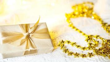 doos met Kerstmis geschenk in licht papier is gebonden met een goud lint Aan wit gebreid achtergrond met decor. gouden slinger van sterren. nieuw jaar. Gefeliciteerd, verrassing. ruimte voor tekst foto