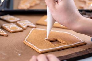 jonge vrouw versiert kerst peperkoek huis koekjes biscuit thuis met glazuur topping in slagroom zak, close-up, levensstijl. foto