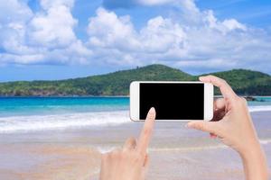 toerist gebruik makend van telefoon Bij de strand met de zee, hand- Holding wit mobiel slim telefoon smartphone, reizen werken concept, wazig achtergrond, dichtbij omhoog. foto