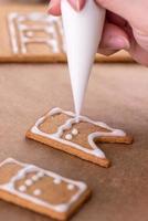 jonge vrouw versiert kerst peperkoek huis koekjes biscuit thuis met glazuur topping in slagroom zak, close-up, levensstijl. foto