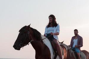 een liefhebbend paar in zomer kleren rijden een paard Aan een zanderig strand Bij zonsondergang. zee en zonsondergang in de achtergrond. selectief focus foto