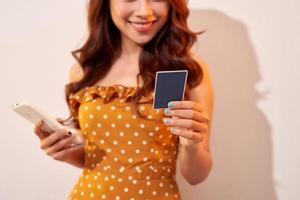 beeld van schattig mooi jong vrouw geïsoleerd over- beige achtergrond tonen plastic credit kaart terwijl Holding mobiel telefoon foto
