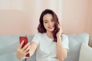 shot van een gelukkige jonge Aziatische vrouw die selfie met haar mobiel neemt terwijl ze in de woonkamer zit. foto