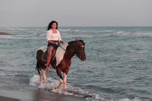 vrouw in zomer kleren geniet rijden een paard Aan een mooi zanderig strand Bij zonsondergang. selectief focus foto