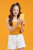 foto van een glimlachen mooi Aziatisch vrouw Holding mobiel telefoon staand over- geel achtergrond.