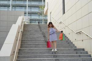 mooi meisje is Holding boodschappen doen Tassen, op zoek Bij camera en glimlachen terwijl wandelen naar beneden de straat foto