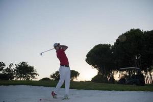 golfspeler raken een zand bunker schot Aan zonsondergang foto