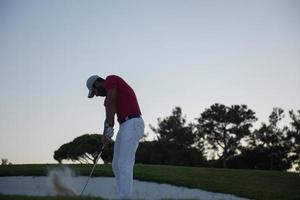 golfspeler raken een zand bunker schot Aan zonsondergang foto