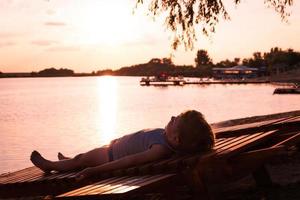 klein jongen ontspannende Aan dek stoel door de water Bij zonsondergang. foto