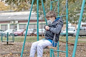 verdrietig weinig jongen met beschermend gezicht masker swinging Bij de park. foto