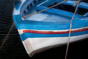 dichtbij omhoog en detail schot van een oud houten visvangst boot geschilderd in blauw, rood en wit verf. de boot staat in de donker water en is gebonden naar de kust met touwen. foto