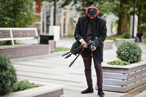 jong professioneel Afrikaanse Amerikaans videograaf Holding professioneel camera met pro apparatuur. afro cameraman vervelend zwart Duraq en gezicht beschermen masker, maken een videos. foto