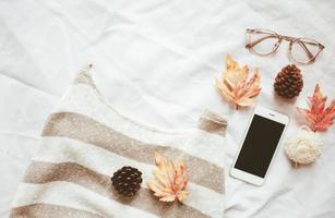 herfst mode stijl concept, trui en smartphone met esdoorn- bladeren Aan wit bed vel achtergrond foto