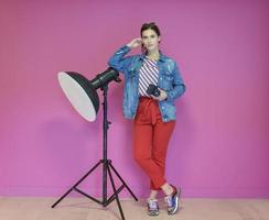 jong vrouw leunend tegen studio zaklamp en Holding camera over- een roze achtergrond foto
