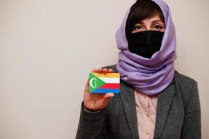 portret van jong moslim vrouw vervelend formeel dragen, beschermen gezicht masker en hijab hoofd sjaal, houden comoren vlag kaart tegen geïsoleerd achtergrond. coronavirus land concept. foto