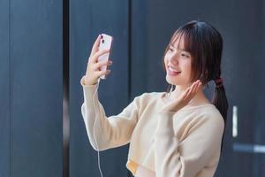 schattig tiener- Aziatisch leerling vervelend een met lange mouwen overhemd is gebruik makend van haar smartphone en hoofdtelefoons naar maken een pret video telefoontje met haar vrienden net zo ze wacht naar invoeren de klas Bij school. foto