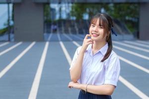 schattig hoog school- Aziatisch leerling meisje in de school- uniform staat en glimlacht gelukkig terwijl vol vertrouwen met de grijs gebouw net zo achtergrond. foto