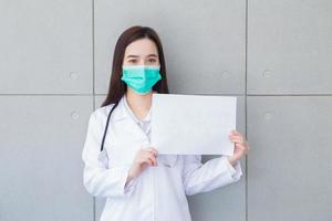 Aziatisch vrouw dokter draagt medisch jas en gezicht masker terwijl shows duidelijk papier wit papier naar Cadeau iets in Gezondheid bescherming concept. foto