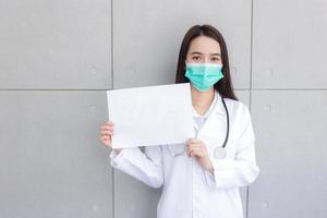 Aziatisch vrouw dokter wie draagt wit medisch jas en gezicht masker staat en shows wit papier naar zeggen iets in coronavirus gezondheidszorg concept. foto
