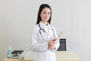 jonge mooie Aziatische vrouw arts werkzaam in het ziekenhuis. ze draagt een wit gewaad en een stethoscoop en houdt een klembord in haar handen. foto