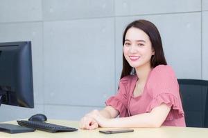 Aziatisch mooi vrouw zit Aan stoel naar werk in kantoor welke heeft computer en mobiel zetten Aan tafel. foto