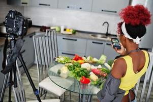 Afrikaanse Amerikaans vrouw filmen haar blog uitzending over gezond voedsel Bij huis keuken en maken afbeelding Aan telefoon. foto