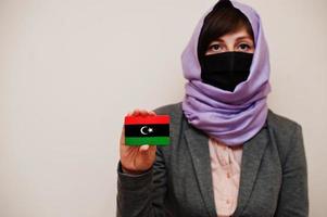 portret van jong moslim vrouw vervelend formeel dragen, beschermen gezicht masker en hijab hoofd sjaal, houden Libië vlag kaart tegen geïsoleerd achtergrond. coronavirus land concept. foto