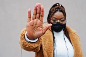 glamoureus Afrikaanse Amerikaans vrouw in warm vacht jas en zwart gezicht masker tonen hou op teken door hand. foto