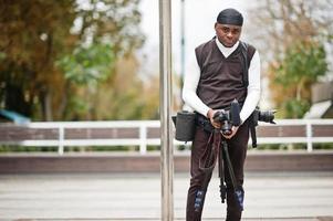 jong professioneel Afrikaanse Amerikaans videograaf Holding professioneel camera met pro apparatuur. afro cameraman vervelend zwart Duraq maken een videos. foto