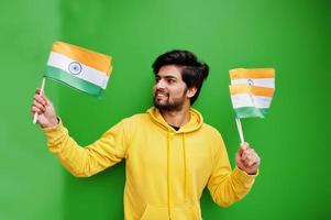 stedelijk jong hipster Indisch Mens in een modieus geel sweater houden vlaggen van Indië in handen. koel zuiden Aziatisch vent slijtage capuchon geïsoleerd Aan groen achtergrond. foto