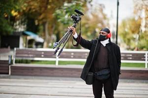 jong professioneel Afrikaanse Amerikaans videograaf Holding professioneel camera met pro apparatuur. afro cameraman vervelend zwart Duraq en gezicht beschermen masker, maken een videos. foto