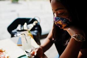 modieus Afrikaanse Amerikaans vrouw, slijtage hand- gemaakt gezicht masker zittend Bij restaurant, kijken Bij mobiel telefoon, terwijl eten pizza. nieuw normaal leven.