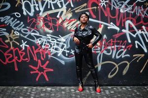 modieus mooi Afrikaanse Amerikaans vrouw poseren in zwart leer jasje en broek Bij straat tegen graffiti muur. foto